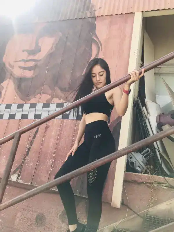 Woman in black athletic wear posing on metal stairs against a corrugated wall with a large face mural.